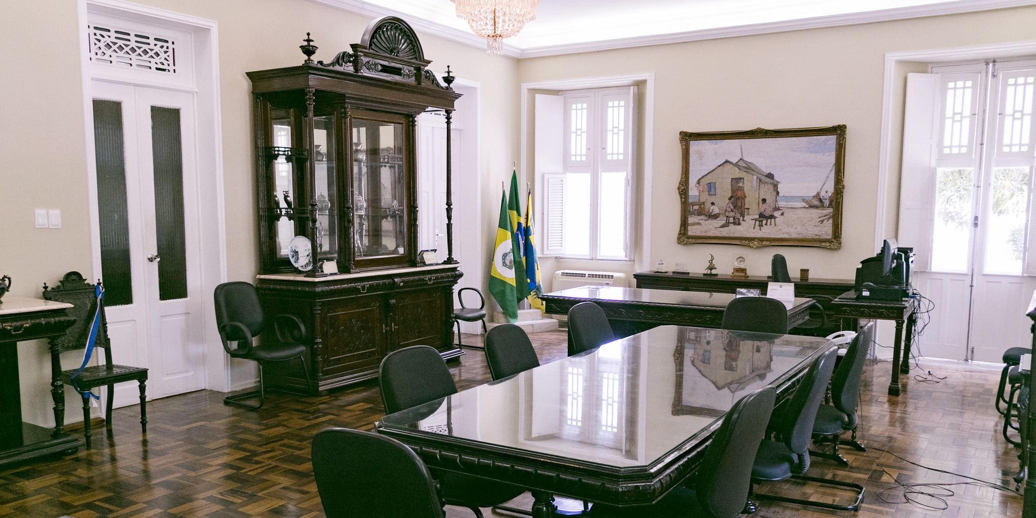 Sala do Gabinete da Reitoria, com piso, mesas e cristaleira em madeira, cadeiras acolchoadas, bandeiras do Ceará e o Brasil, quadro do pintor Raimundo Cela e equipamentos de escritório