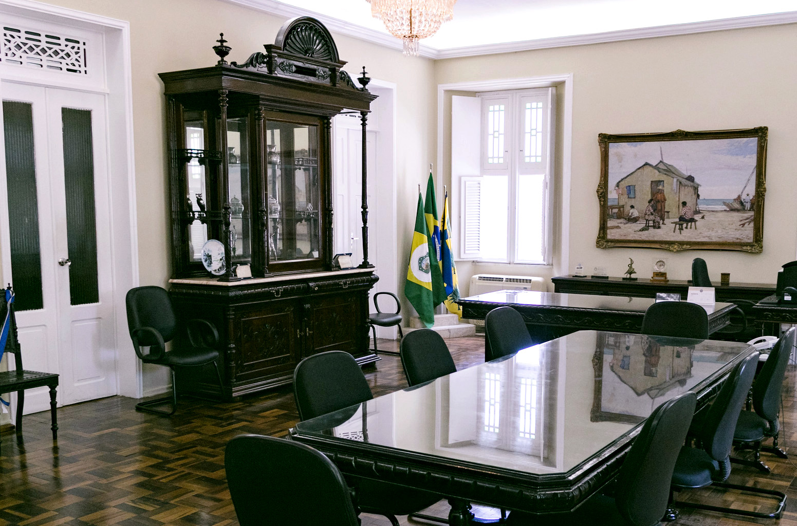 Sala do Gabinete da Reitoria, com piso, mesas e cristaleira em madeira, cadeiras acolchoadas, bandeiras do Ceará e o Brasil, quadro do pintor Raimundo Cela e equipamentos de escritório